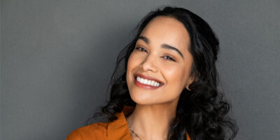 Smiling woman with long dark hair and orange top against gray background.